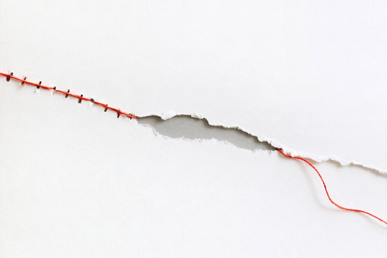 A torn sheet of white paper being sewn back together with orange thread